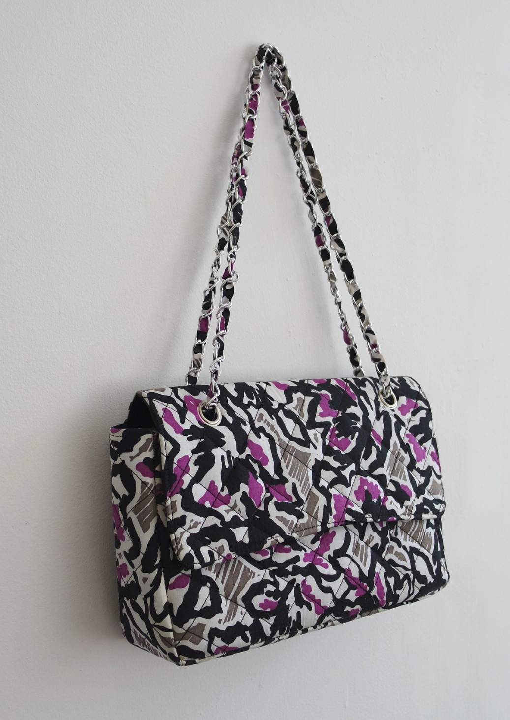 An Ankara African print handbag in hues of grey, purple and black. The bag has a chain handle detailing and fold over flap.