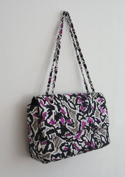 An Ankara African print handbag in hues of grey, purple and black. The bag has a chain handle detailing and fold over flap.